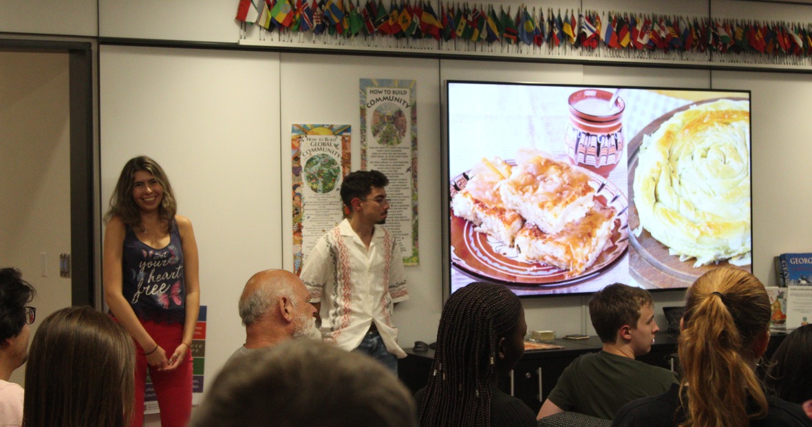 Two students smile as they present. A TV screen displays Bulgarian food.